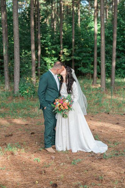 A newlywed couple kissing while standing in a wooded area
