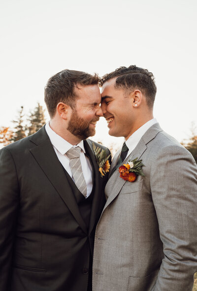 A male couple touches noses and foreheads as they laugh together on their wedding day. wearing suits and fall floral boutonnieres.