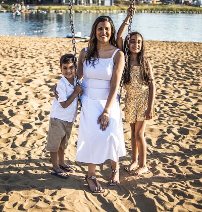 Mother in a white dress sits on a playground swing while her son and daughter stand next to her 
