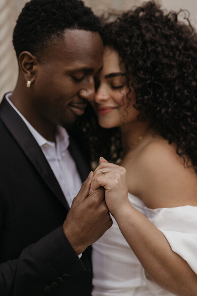 A tender moment between a newlywed couple, with the bride, in a white dress, resting her head on the groom's shoulder while he gently holds her hand, both smiling softly with eyes closed