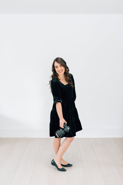 Wedding photographer Sarah Noel smiles while holding her Canon R6 camera, wearing a black velvet dress and black flats at a photography studio