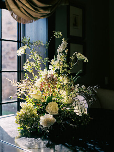 An elegant wedding floral centerpiece placed next to a window in a darker room at Capitol City Club. The florals look like they're glowing from the light coming from the window. 