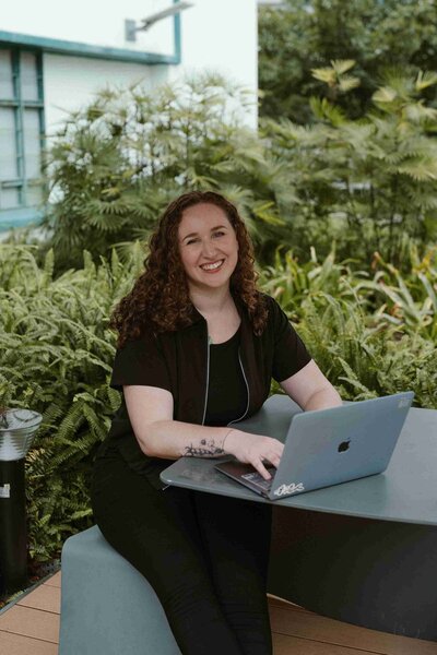 Woman with curly hair smiles while typing on a laptop at an outdoor table, surrounded by lush greenery. The atmosphere is relaxed and cheerful.