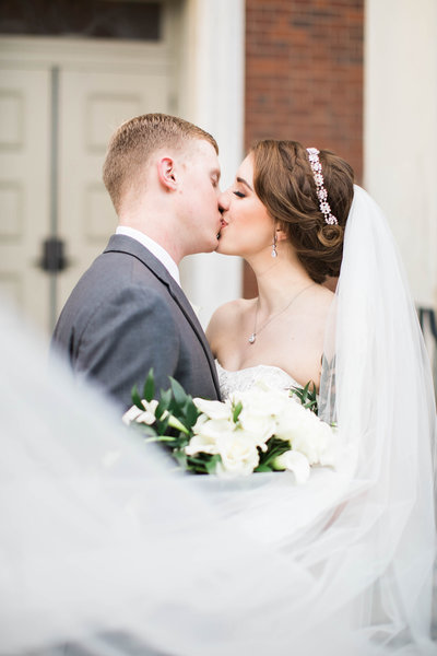 bride and groom in wedding attire with a long cathedral veil surrounding them kissing outside of a church.