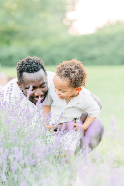 A dad crouches down to smell a lavender bush with his toddler son