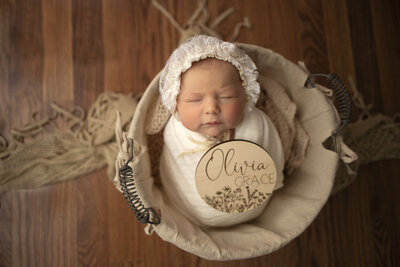 Swaddled baby wearing a bonnet sits in a basket with her name engraved on a plaque.