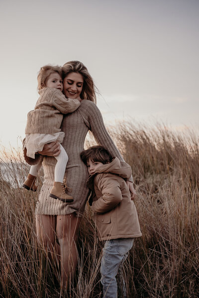 A woman and a child stand together on rocky terrain with the sun setting behind them, casting a warm glow. The woman is wearing a dark dress, and the child is in a white dress and sneakers. This moment, captured by Wollongong family photography, beautifully encapsulates the essence of natural family photography in NSW.