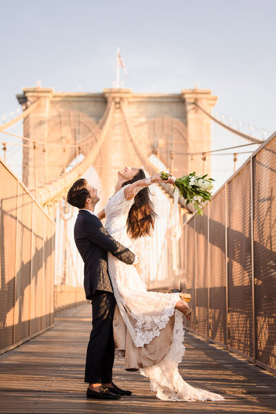 Groom lifts bride in celebration on the Brooklyn Bridge at sunrise, her lace gown flowing as she holds her bouquet high, captured by NYC wedding photographer Heritage House.
