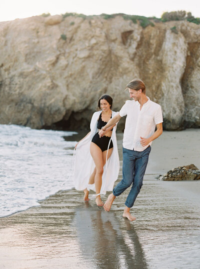 woman in black bikini holding hands with man in white shirt walking along the edge of the beach with cliffs behind them