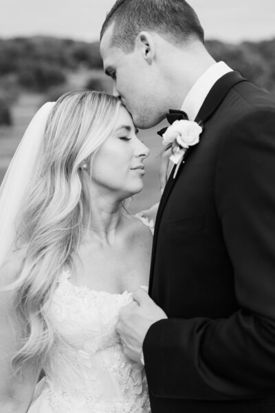 A groom in a black tux kisses his bride on the forehead