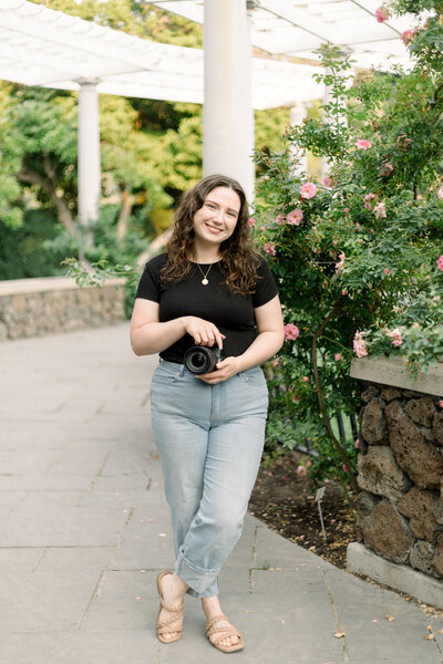 female photographer holding camera and smiling at camera lens taken by spokane wedding photograpeher