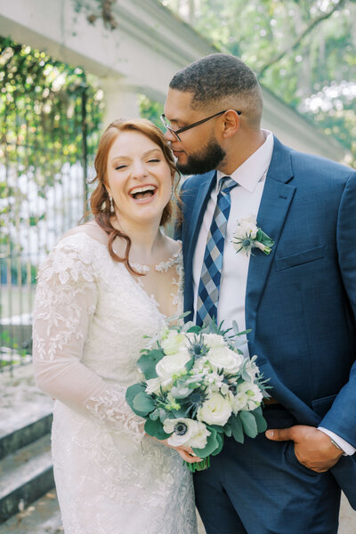 Groom kissing his laughing bride on the temple while holding a white and green bouquet during their elopement in Savannah, GA.