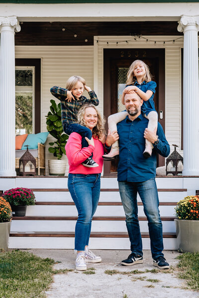 A family of four stands proudly on the steps of their porch. The children sit on the parents' shoulders