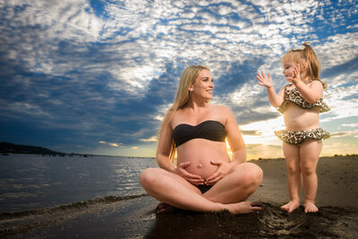 A beautiful maternity photo capturing a pregnant mother in a bikini top sitting on the beach, smiling at her young child in a leopard print swimsuit, against a dramatic sunset sky and calm water