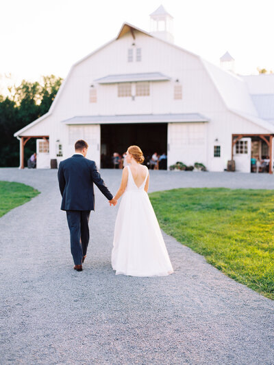 Bride and groom walking and holding hands in front of the Wildflower On Watts white barn venue