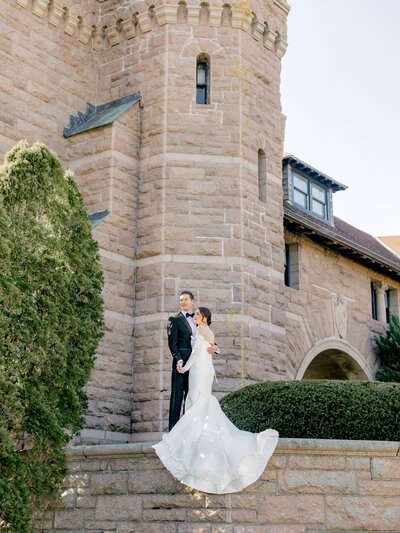 Bride and groom, holding hands, standing on a stone wall ledge with Oceancliff Hotel behind them.