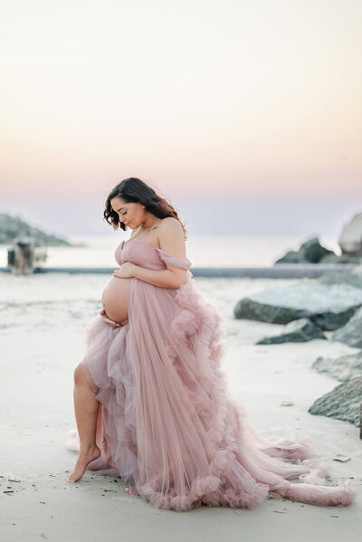 A mama to be looks lovingly down at her her bump while wearing a pink tulle gown on the beach.