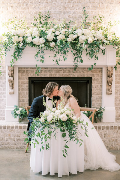 A bride and groom kiss at their floral adorned sweetheart table during their reception at Harvest Hollow by Huntsville wedding photographer, Kelsey Dawn Photography