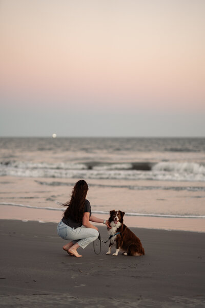 Girl bent down petting her dog on the beach. girl is looking at the ocean during sunset and the dog is looking at the camera.