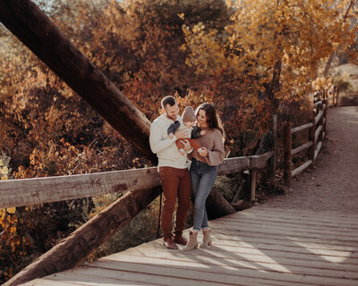 A young family with one 6 month old baby boy stand on a bridge at Sunset in the fall in Colorado interacting with each other, it's such a beautiful location that it looks like a magazine cover.