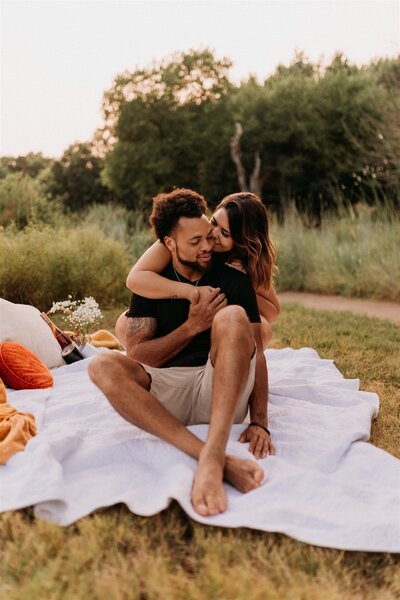 Girl holds onto guy sitting on picnic blanket and kisses his cheek