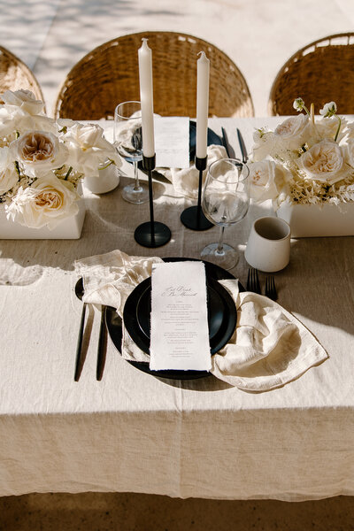 Close-up of boho wedding table design in Mexico with modern black place settings, ivory roses, and taper candles.