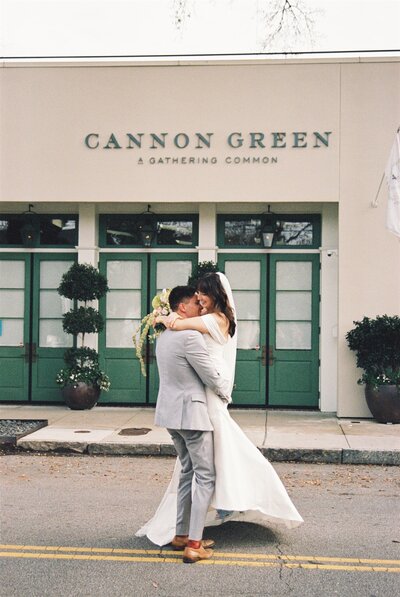 A groom is picking up his bride and twirling her around in front of their wedding venue Cannon Green in Charleston