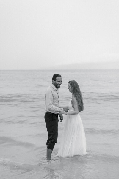 Couple holding hands and laughing in ankle deep water wearing their wedding clothes photographed on black and white film