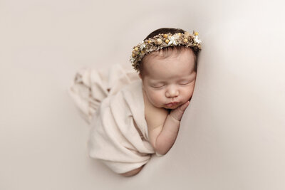Newborn girl sleeps with hand under chin in a cream wrap with flower halo.