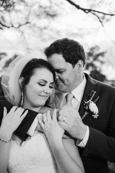 A groom wraps his arms around his bride in a tender embrace, pressing a loving kiss to her temple as she smiles softly. Captured in timeless black and white, this intimate moment reflects the deep connection and joy of their wedding day, beautifully documented by an Alabama wedding photographer.