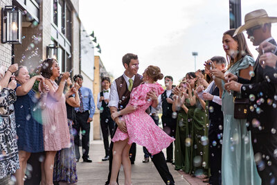 A couple dances joyfully in the center of a cheering crowd outdoors, surrounded by people blowing bubbles and clapping, dressed in semi-formal attire on a bright day.