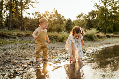 Toddler boy watching his sister play in the shallow lake water. 