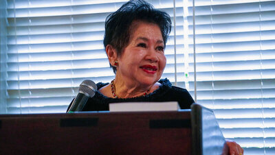 Nita Tin speaking at an event in Tennessee, smiling warmly while standing at a podium with white blinds in the background. Wearing black with ruby earrings and necklace, her expression shows affection and care.