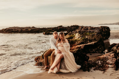Expecting couple sitting on rocks at the beach during a golden-hour maternity photoshoot in Laguna Beach, California, captured by Southern California photographer Brittani Luce