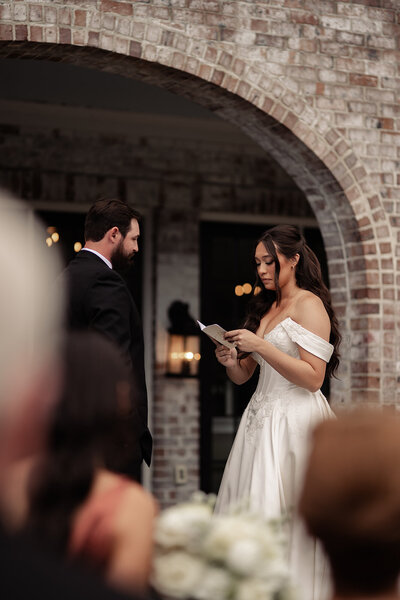 Bride speaking her vows to her husband with a soft face and vow book in hand. The outline of her family in the forground.