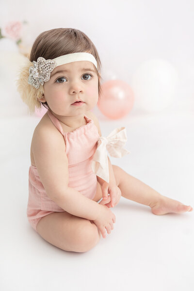 1st birthday boho Gatsby themed studio portrait of  a baby girl with large brown eyes wearing a peach onesie and a big cream bow and a cream feathered and jeweled headband with peach balloons and flowers in the background