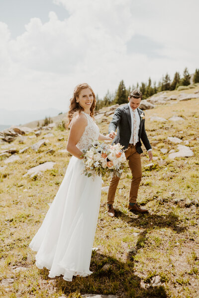 Bride hold flowers stands on a hill with groom in the background