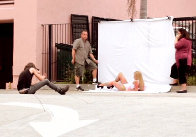 Behind the scenes musician photoshooot Savannah Lynx lying in white bathing suit with painted hand prints against white backdrop set up in alley Mark Maryanovich taking her picture