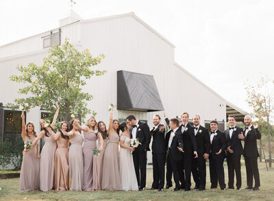 Bridal party in front of white barn wedding venue called Dream Point Ranch with bridesmaids wearing blush pink bridesmaid dresses and groomsmen wearing black tuxedos with bride and groom kissing and the wedding party cheering and smiling and photography by Tulsa Wedding Photographer Laura Eddy