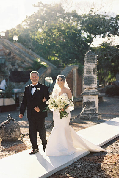 Bride walking down the aisle with her father, holding a white rose bouquet at a destination wedding in Altos de Chavón — photographed by Asia Pimentel.