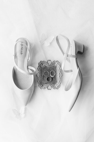Black and white photo of wedding shoes with rings on a tray in between the shoes
