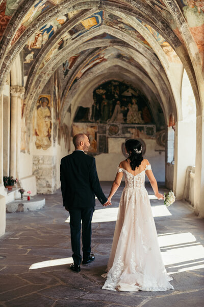 The bride and groom walk down the fresco-adorned abbey in Bressanone, surrounded by historic charm and artistic beauty, creating a timeless and magical moment