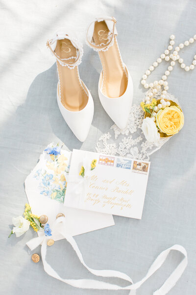 Flat lay photo of wedding stationery, bridal heels, jewelry and florals