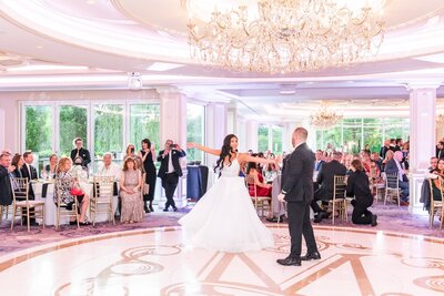 The bride glides across the floor in a ballgown-style wedding dress, arms outstretched as the groom leads their first dance. The grand reception space, with floor-to-ceiling windows and opulent chandeliers, is filled with the glow of candlelight and the joyful gaze of wedding guests