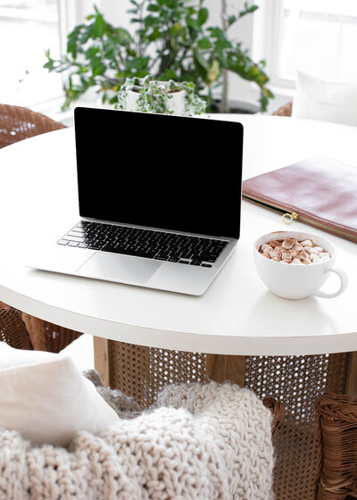 A work from home setup with an open laptop on a table next to a cup of hot chocolate.