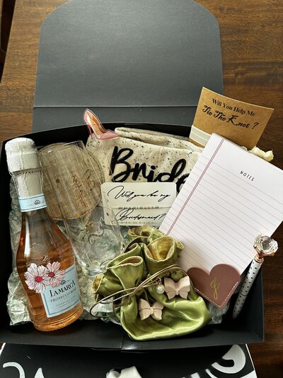A box filled with special gifts for the bride's maids to-be. A bottle of pink prosecco adorned with pretty pink flowers in the upper left hand corner. overlapping the lavel. An engraved stemless campagne glass with the name Jamie hand engraved across the front. A round metalic rose gold  coaster. A set of pink butterfly shaped champagne fizzies.  A pair of socks that say "bride's" on the sole of one and "maid" on the sole of the other.  A pink note pad and white polka dot pen with a big crystal diamond on top.  A mauver colored heart shaped corner bookmark with the letter J foiled in gold on it. and a beaded morse code bracelet that says "bridesmaid". the tiny seed beads are pink and green with white beads for spacers.