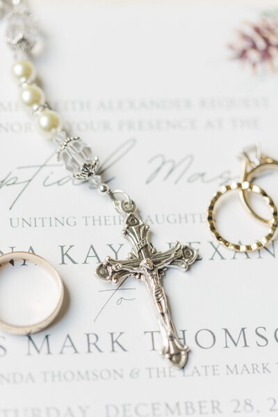close up of Catholic rosary beads with a cross and wedding rings laying on top of a wedding invitation
