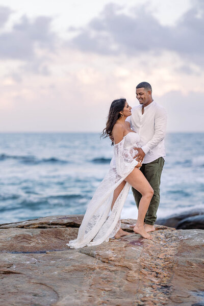 breathtaking maternity photo of woman and man facing one another and smiling on a cliff overlooking the ocean in Narragansett Rhode Island