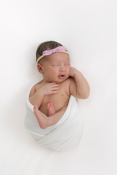 Newborn baby laying on a white backdrop wrapped in white fabric with a purple bow on her head as she sleeps on her back