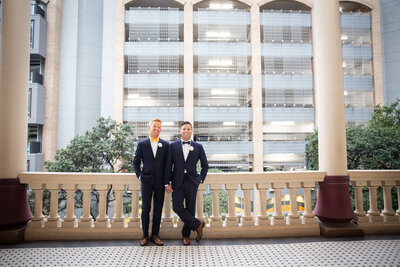 Austin-based wedding photographer capturing two grooms standing on a balcony in front of a building.